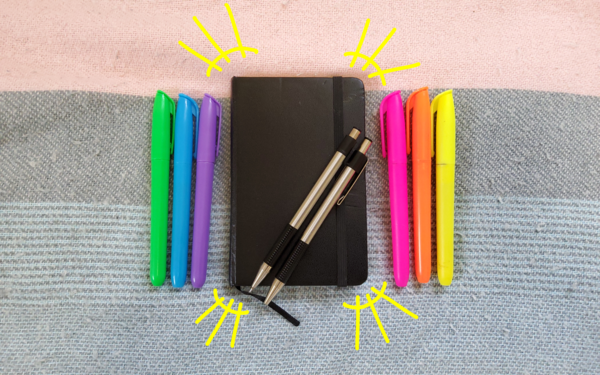 [ID: another photo of tiny notebook, but with highlighters lined up on both sides of it. Two pens have been laid across it and yellow solrads have been drawn around it. End ID.]